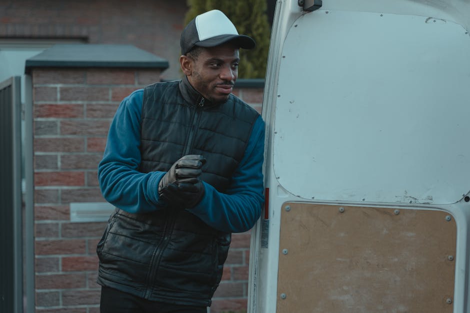 A young man with a beard, wearing a black cap and a white polo shirt, stands inside the open back of a moving van loaded with brown cardboard boxes of various sizes, some wrapped in plastic or packing material. The van is parked on a paved area, with the side door open revealing the interior cluttered with boxes on the floor and stacked towards the ceiling. The man has a serious expression, arms crossed, as he supervises the home relocation process, with visible equipment such as straps and blankets used for furniture transport. Natural daylight illuminates the scene, highlighting the packing and loading activities involved in professional house removals, as managed by [COMPANY_NAME] for stress-free moving services. The environment outside the van suggests a residential area, supporting the context of packing and logistics related to house moves, with the scene emphasizing the careful handling and transportation of possessions during a furniture transport operation.