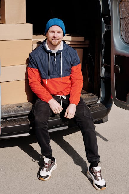 A young man sitting on the open rear door of a black moving van parked outside a residential building. Inside the van, several large cardboard boxes and flat-packed furniture pieces are visible, secured with protective plastic wrap and foam padding. The man is dressed in a blue beanie, a navy and red waterproof jacket, black pants, and white sneakers. He is smiling and looking towards the camera, with one hand resting on his knee. The scene captures the loading process during a home relocation, with the man appearing to be taking a break or preparing to continue loading. The environment is well-lit, and the pavement beneath the van has some shadow refraction from the vehicle, indicating daylight. This image relates to furniture transport and packing during property removals, illustrating the logistical aspect of house removals with a focus on the careful handling of packed items during a moving service managed by Man with Van Highbury, ensuring efficient and professional logistics for clients with difficult access points.