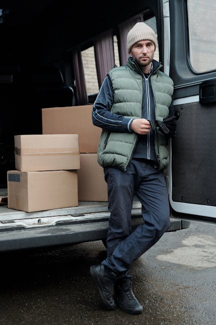 A man wearing a white T-shirt, jeans, and a black cap is assisting with a home relocation, carrying three cardboard boxes stacked together while standing beside a white panel van parked on a residential street. The van, part of the services offered by Man with Van Highbury, is positioned next to the pavement in front of a suburban house with a pitched roof, multiple skylights, and a balcony. The rear doors of the van are open, with additional cardboard boxes placed on the ground near the vehicle. Inside the house, visible through the doorway, is a clutter-free entrance area, indicating the start of a furniture transport and packing process. The scene captures the loading process involved in difficult access removals on the Highbury Estate, with the man preparing to load or unload the boxes from the van, supported by moving equipment like trolleys or straps that might be present nearby. Bright daylight and neatly maintained greenery and fencing establish an organized and professional atmosphere for house removals conducted by Man with Van Highbury.