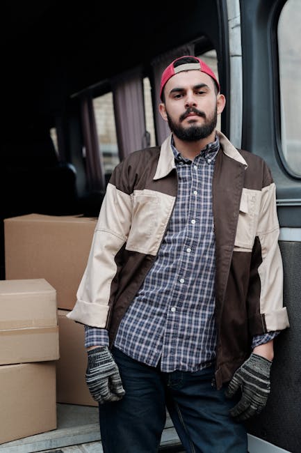 A man with a beard, wearing a beige and brown jacket over a checked shirt, black gloves, and a backwards red cap, standing inside a van used for home relocation. He is positioned near the open rear door of the vehicle, with cardboard boxes stacked behind him on the van's floor, prepared for furniture transport and packing during a house move. The van's interior is lit naturally, and the background shows an urban environment through the window. This scene captures the loading process associated with professional removals and moving services, as provided by Man with Van Highbury, in alignment with house removal logistics and packing activities.
