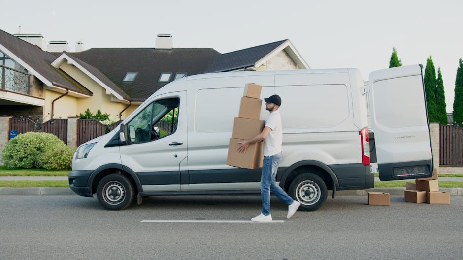 A man with a beard, wearing a beige and brown jacket over a checked shirt, black gloves, and a backwards red cap, standing inside a van used for home relocation. He is positioned near the open rear door of the vehicle, with cardboard boxes stacked behind him on the van's floor, prepared for furniture transport and packing during a house move. The van's interior is lit naturally, and the background shows an urban environment through the window. This scene captures the loading process associated with professional removals and moving services, as provided by Man with Van Highbury, in alignment with house removal logistics and packing activities.