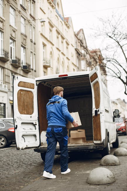 Two movers from Man with Van Highbury are engaged in the loading process of a van positioned on a paved street, with its rear doors wide open. They are lifting and carrying a large, rectangular piece 