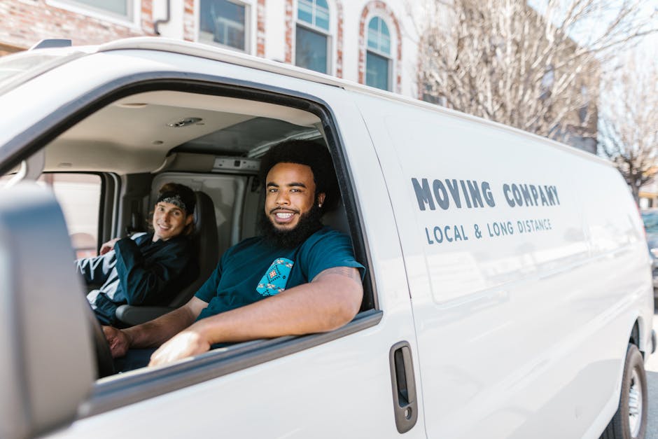 Inside a white moving van with the side door open, two movers from Man with Van Highbury are seated and smiling. The man in the foreground has a dark beard, curly hair, and is wearing a navy blue t-shirt with a logo, while the woman behind him has long dark hair, a black headband, and is dressed in a dark jacket. The van's interior reveals packed cardboard boxes, furniture blankets, and plastic wrap, indicative of a home relocation process. The vehicle is parked on a street with buildings and leafless trees visible in the background, suggesting an urban setting. The scene captures the loading or unloading phase of furniture transport and packing for a move, aligned with regulations for parking during removals in Islington as detailed on the webpage. Man with Van Highbury provides local and long-distance moving services, supported by careful handling of household items, equipment like trolleys and straps, and efficient loading procedures.