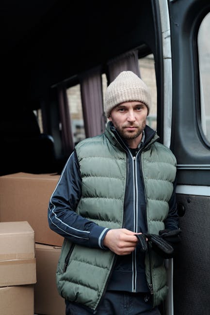 A man with a serious expression is standing inside a vehicle used for home relocation, holding a black strap or tie in his hands. He is dressed warmly with a light grey knitted beanie and a padded green and navy blue jacket, indicating preparations for moving activities. Behind him, there are cardboard boxes stacked on the vehicle's interior, some covered with protective wrapping. The vehicle appears to be a van, with a dark interior and a sliding door open, revealing the boxes and a glimpse of the environment outside. The scene captures part of the loading process managed by Man with Van Highbury, a professional service specializing in furniture transport, packing, and moving logistics. The lighting inside the van is natural, suggesting daytime, and the setting emphasizes the practical steps involved in a house removal or furniture transport task.