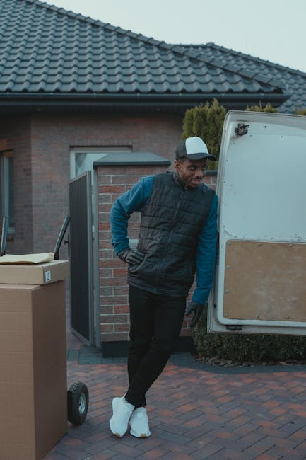 A man dressed in a blue long-sleeved shirt, black trousers, and a black puffer vest, wearing a grey cap and white sneakers, stands beside an open moving van parked in front of a residential building with a brick exterior and a tiled roof. Inside or near the van, a large cardboard box is visible on a hand trolley, indicating a home relocation process. The man appears to be taking a break during the furniture transport and packing and moving activities associated with house removals. The setting is outdoors on a paved driveway with some greenery and the open garage door of the house in the background. The scene captures the staging of loading or unloading household items as part of professional removals services provided by Man with Van Highbury.