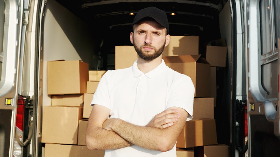 A man with a serious expression is standing inside a vehicle used for home relocation, holding a black strap or tie in his hands. He is dressed warmly with a light grey knitted beanie and a padded green and navy blue jacket, indicating preparations for moving activities. Behind him, there are cardboard boxes stacked on the vehicle's interior, some covered with protective wrapping. The vehicle appears to be a van, with a dark interior and a sliding door open, revealing the boxes and a glimpse of the environment outside. The scene captures part of the loading process managed by Man with Van Highbury, a professional service specializing in furniture transport, packing, and moving logistics. The lighting inside the van is natural, suggesting daytime, and the setting emphasizes the practical steps involved in a house removal or furniture transport task.