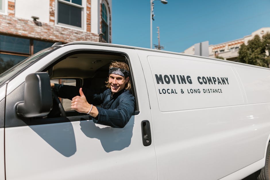 A smiling male mover with long hair, wearing a black bandana and navy-blue shirt, is sitting in the driver's seat of a white moving van with the door open. He is giving a thumbs-up gesture and looking towards the camera. The van has the text 'MOVING COMPANY LOCAL & LONG DISTANCE' printed on its side. In the background, there are modern buildings with brick facades and glass windows, along with a clear blue sky and some trees. The scene is outdoors, likely on a residential street, during daytime. This image illustrates the loading process and professional home relocation services provided by Man with Van Highbury, showcasing a vehicle ready for furniture transport and packing during a move from Highbury Grove.
