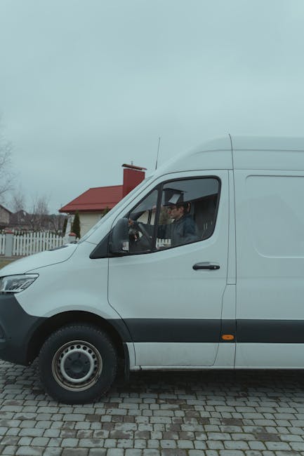 A white commercial van parked on a cobblestone driveway in a residential area during daytime, with the driver visible through the open window. The driver, wearing a dark jacket and glasses, appears to be preparing for a home relocation or furniture transport as part of a house removal service. In the background, a red-roofed house, a wooden fence, and some trees are visible under a cloudy sky. The scene reflects typical packing and loading activities carried out during moving processes managed by Man with Van Highbury, aligning with the topics of removals and moving logistics.