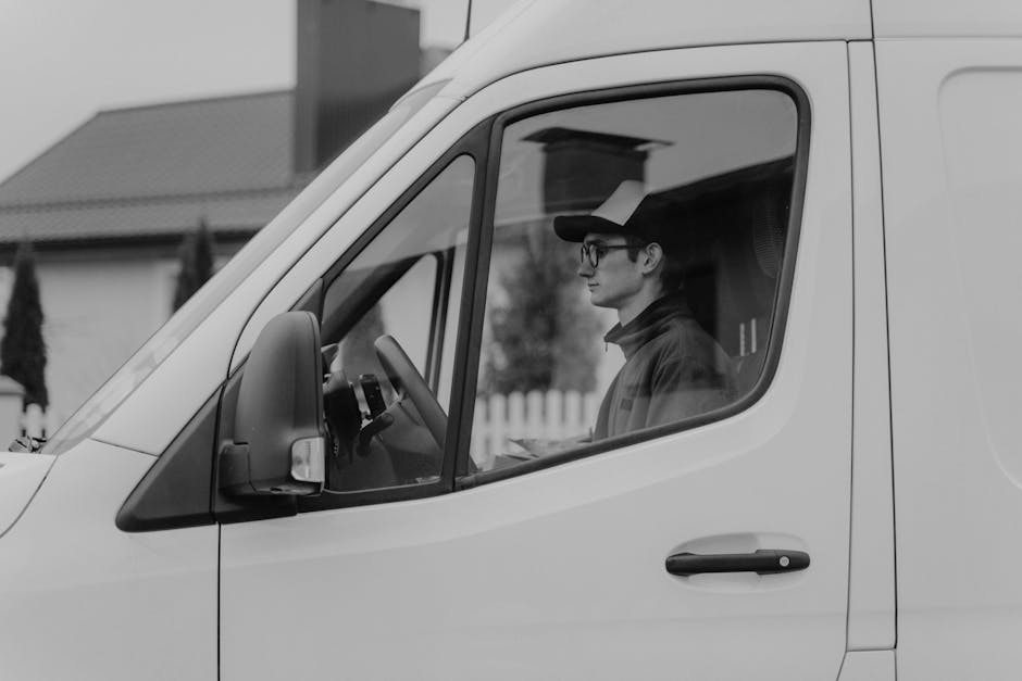 A man wearing glasses and a cap is seated inside a white van, with the side door open for loading or unloading. The van is parked on a residential pavement, with a house and a garden fence visible in the background. The interior of the van shows some packaging materials and boxes, indicating a home relocation process. The man appears to be reviewing documents or instructions related to the moving or transportation task. The scene captures the loading process typical of house removals, facilitated by a professional moving service such as Man with Van Highbury. The environment is illuminated by natural daylight, and the photograph emphasizes the vehicle's role in furniture transport and packing and moving activities.