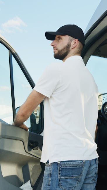 A man wearing a white t-shirt, black cap, and blue jeans is seated on the ground outside a white van with an open rear door, which is filled with numerous packed cardboard boxes of various sizes, some